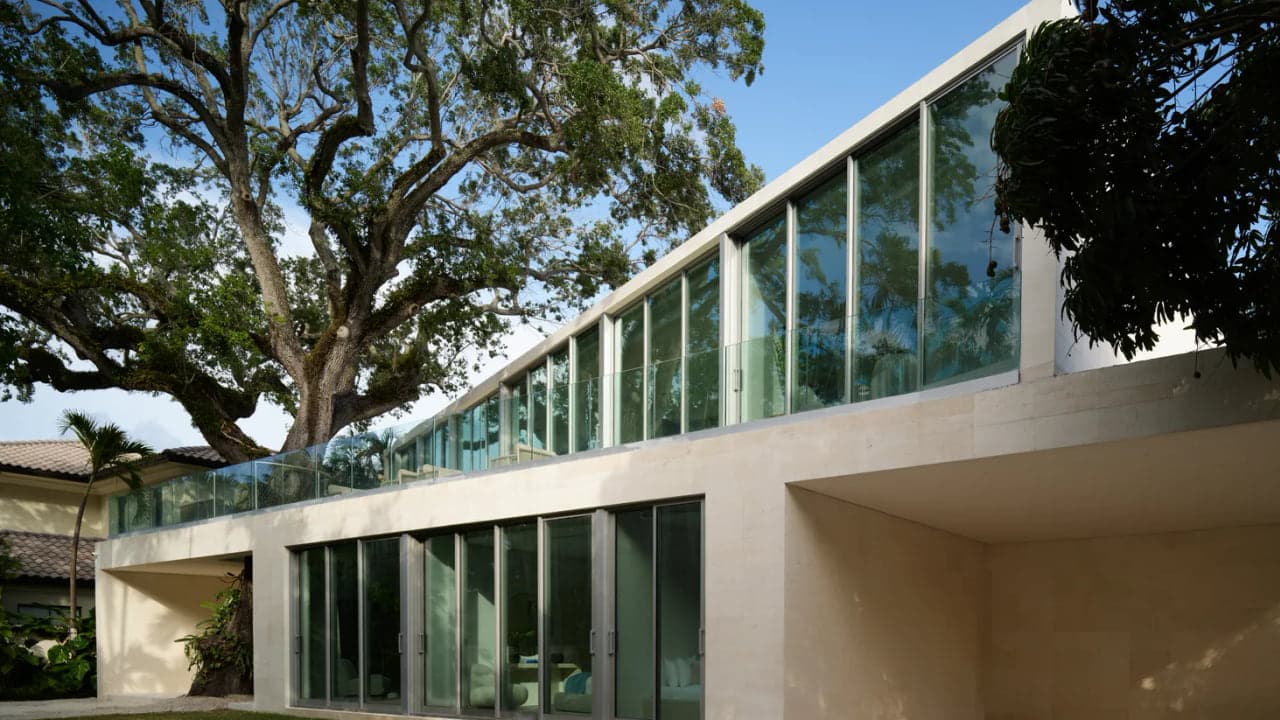 Cantilevered glass and concrete volume of a modern residence extending over a ground floor beneath a mature oak canopy, showing structure carrying a room without a column underneath