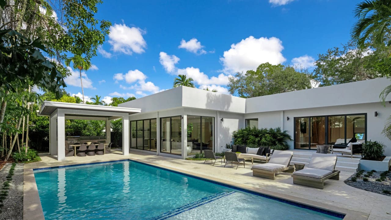 A modern Miami house with deep roof overhangs, floor to ceiling glass and a pool in bright midday sun, no people in frame