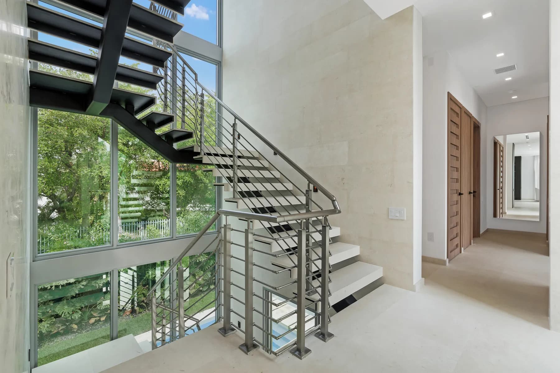 Open riser staircase with pale treads on dark steel stringers and a stainless steel horizontal bar railing beside a double height window in the Crystal residence by LUXOM, Miami