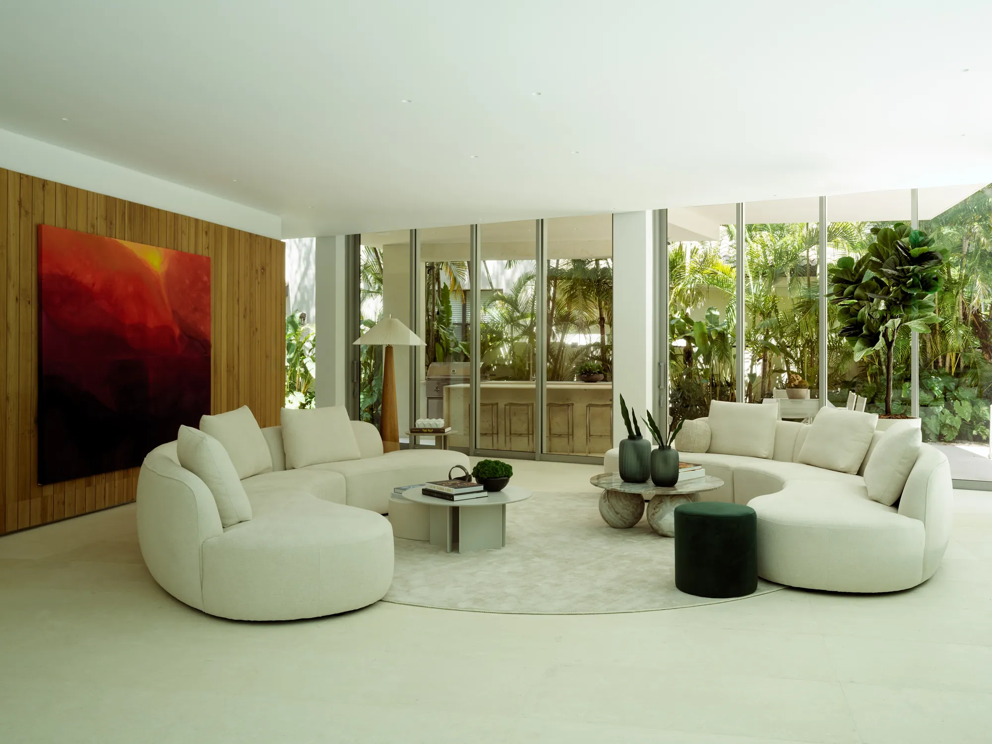 Finished Onaway living room in Coconut Grove: a large area rug under curved cream sofas, stone coffee tables, a cone floor lamp, potted plants, and a large abstract red and orange painting on the wood paneled wall