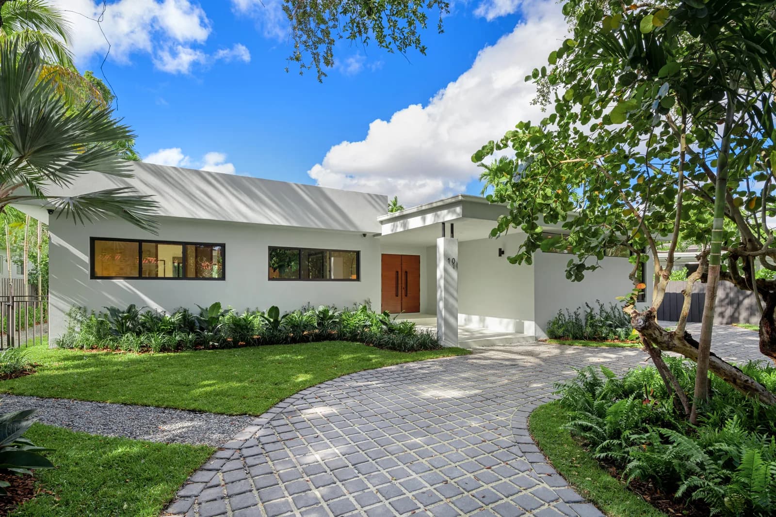 Curved gray paver driveway leading through lawn, ferns and a mature tree to a white single story residence with a wood front door in the Pinta residence by LUXOM, Miami