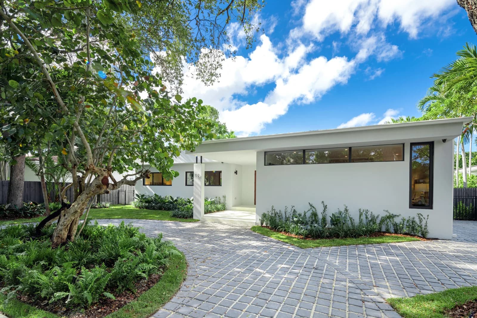 Single story white residence with a mono pitch shed roof over a band of clerestory windows, seen across a paver motor court with a mature tree in the Pinta residence by LUXOM, Miami