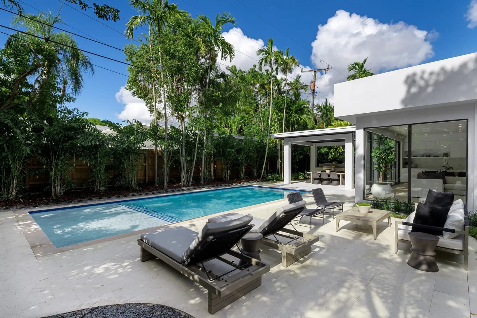 Rectangular pool with a pale paved deck, loungers and a covered pavilion under tall palms in the Pinta residence by LUXOM, Miami