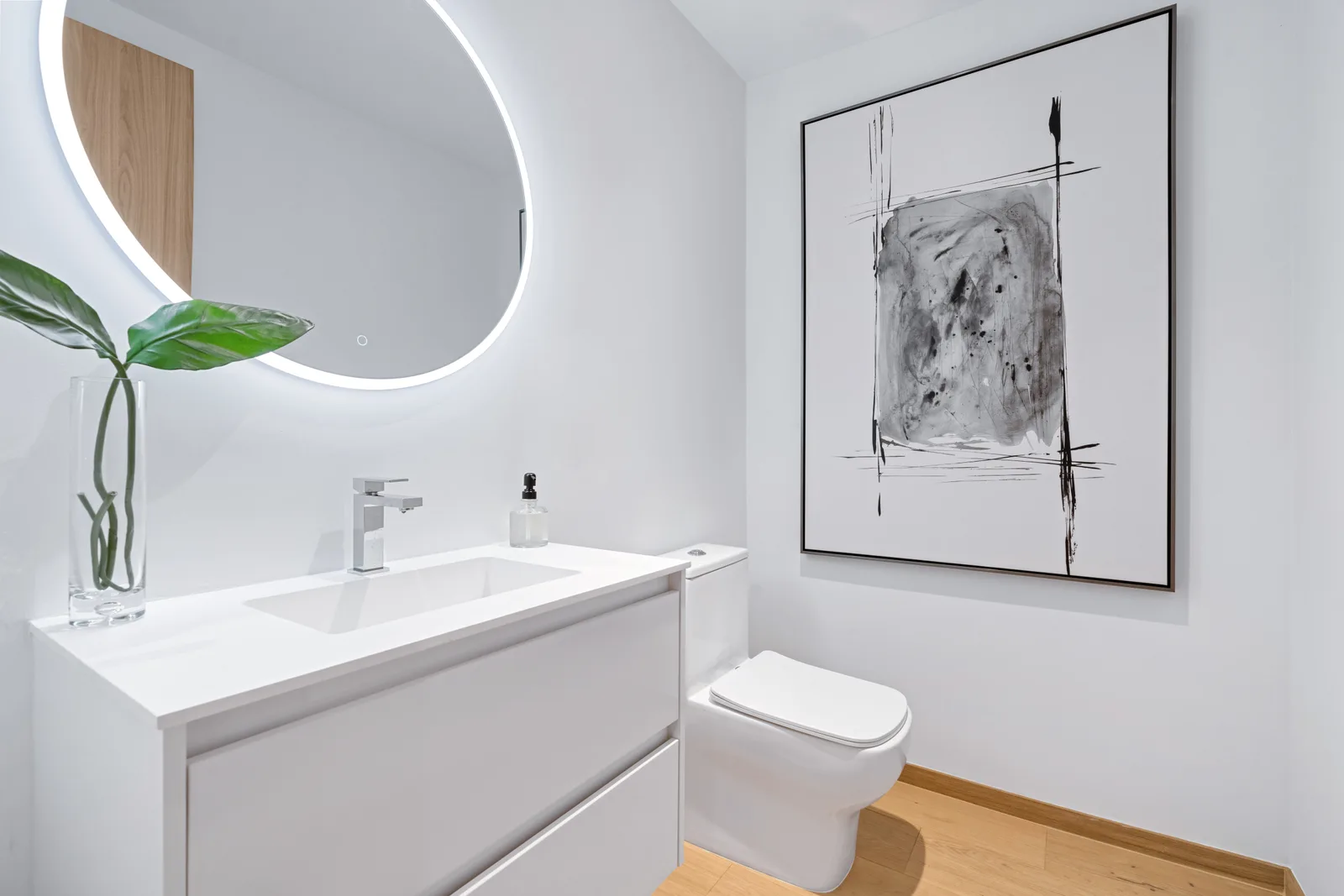Powder room with a white two drawer vanity and integrated basin, a single lever faucet and a backlit round mirror beside a framed abstract artwork, in the Pinta residence by LUXOM, Miami