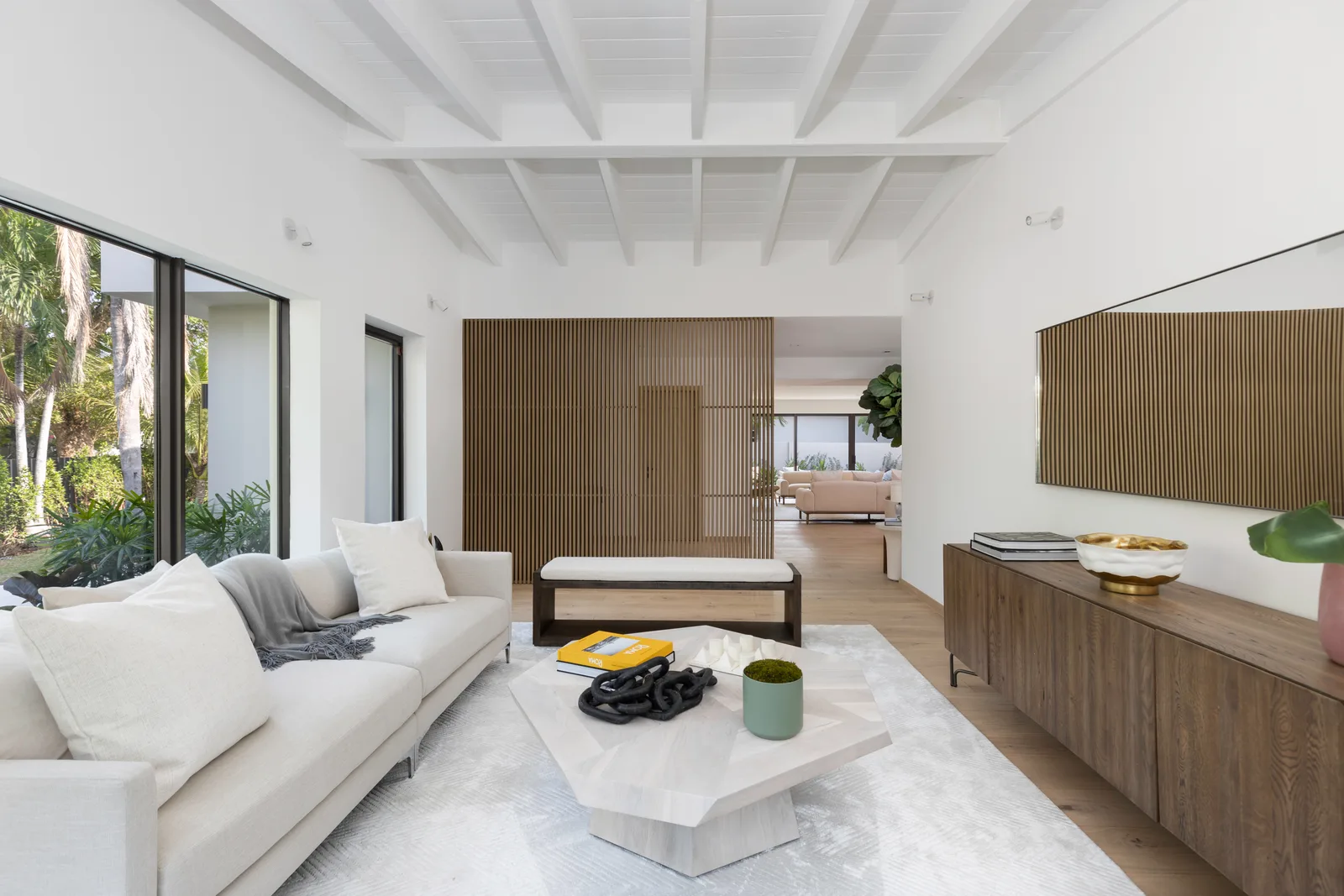 Living room with a white sofa, a faceted white coffee table, a wood slat feature wall and a white beamed ceiling, with sliding glass doors to the garden, in the Samana residence by LUXOM, Miami