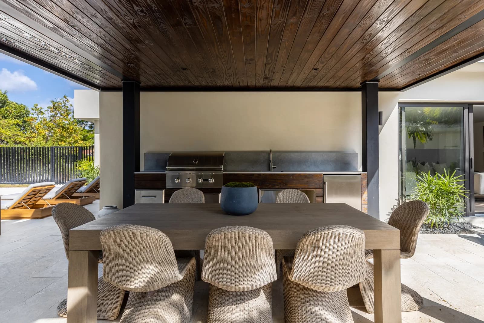 Covered outdoor kitchen with a built in grill and sink under a dark wood ceiling, a wood dining table with woven chairs, and pool loungers beyond, in the Samana residence by LUXOM, Miami