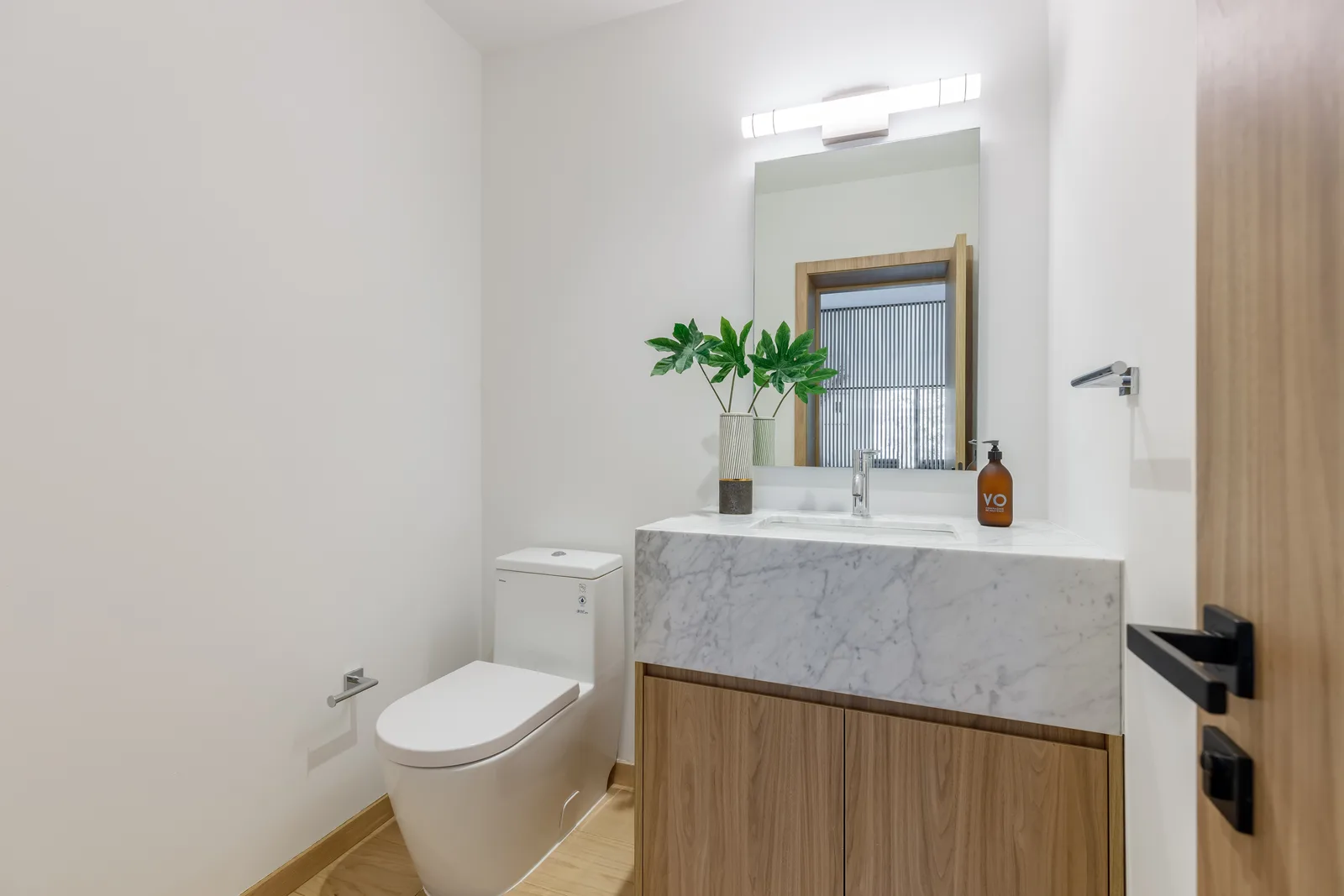 Powder room with a marble clad vanity over wood doors, a chrome faucet and towel ring, and a matte black lever handle on the wood entry door, in the Samana residence by LUXOM, Miami