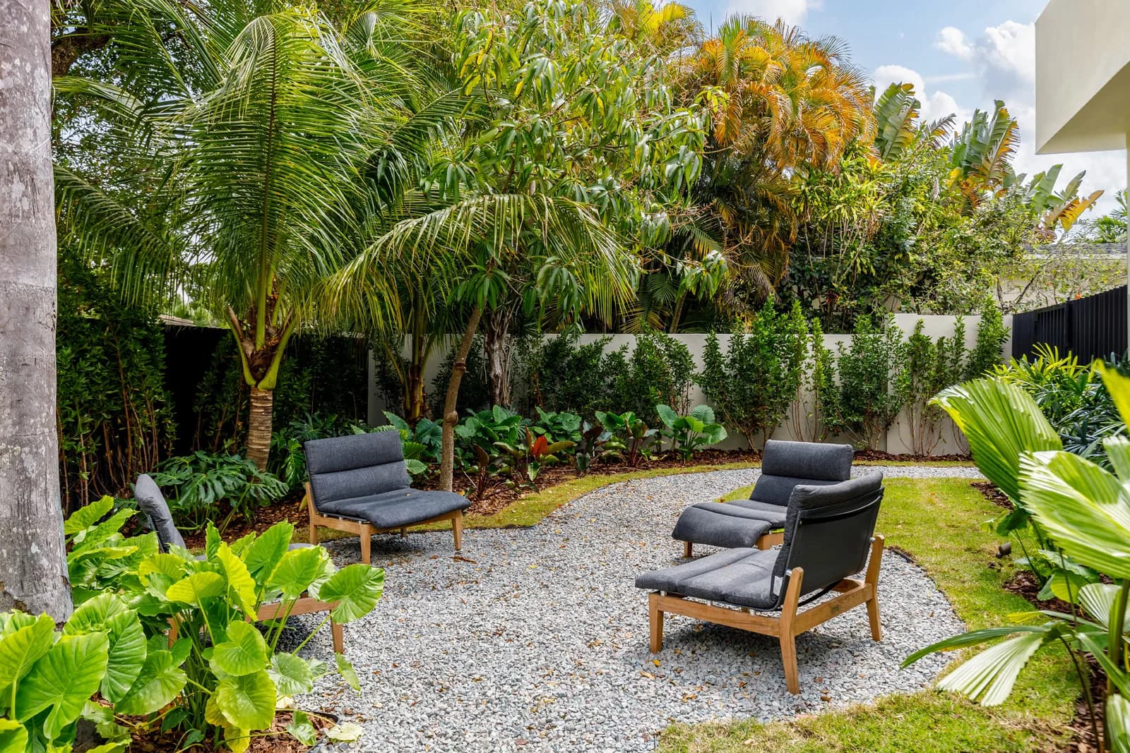 Gravel garden with four lounge chairs under tall palms, framed by layered tropical planting and a hedge along the boundary, in the Samana residence by LUXOM, Miami