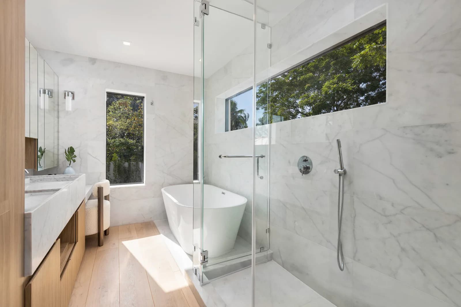 Marble-clad bathroom with book-matched veining on the shower wall, a soaking tub and a garden-view window in the Samana Residence by LUXOM, Miami